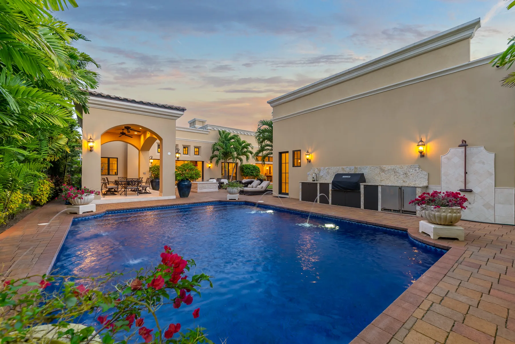 An outdoor swimming pool beside a covered patio, with tropical plants around the edges.