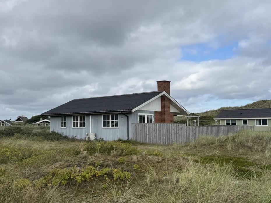 Una casa baja con techo oscuro se ve tras una cerca desgastada, rodeada de hierba.