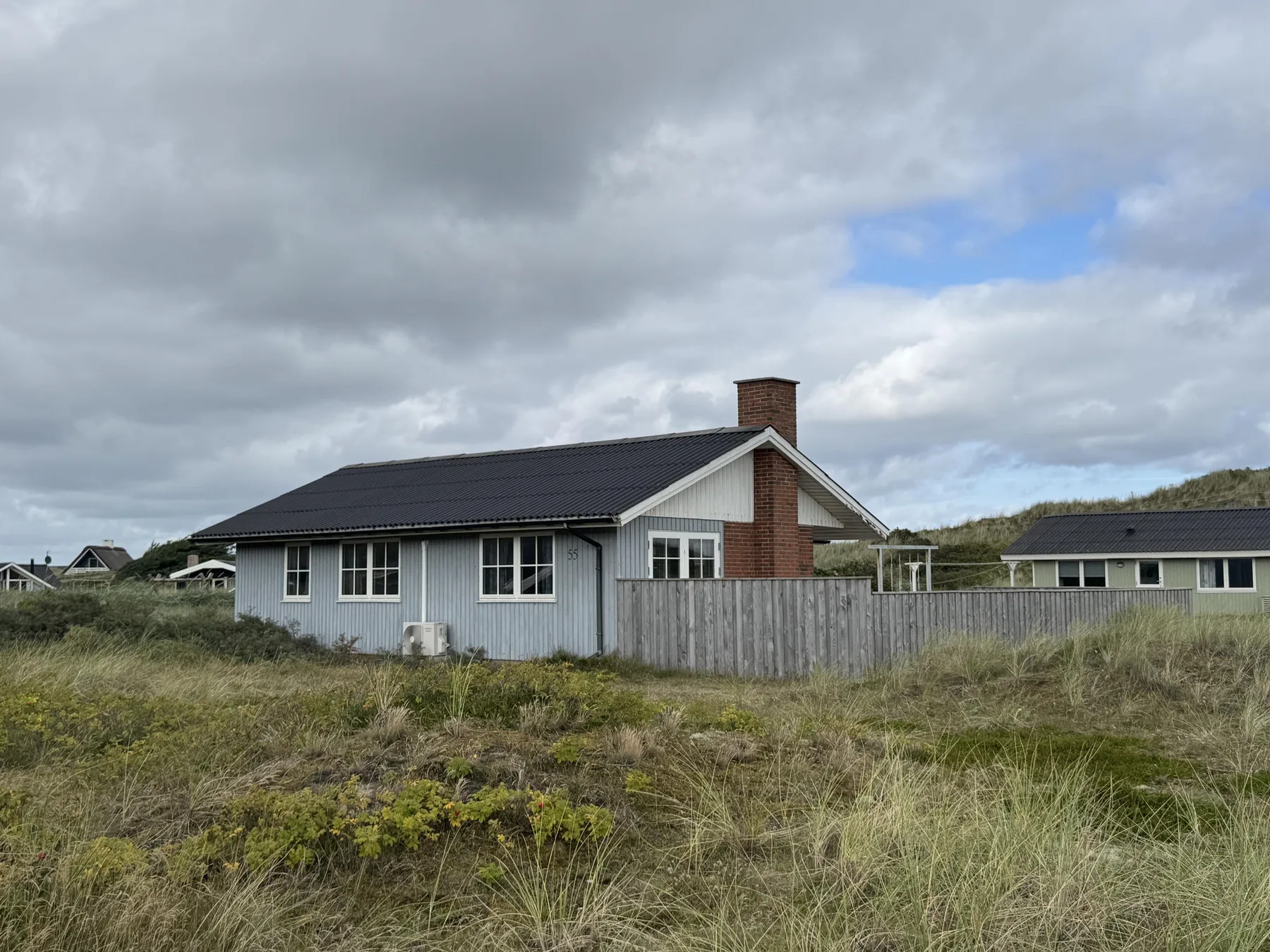 Una casa baja con techo oscuro se alza tras dunas cubiertas de hierba bajo un cielo nublado.