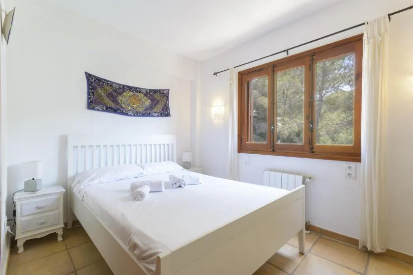 A bright bedroom with a white bed beside large wooden-framed windows.