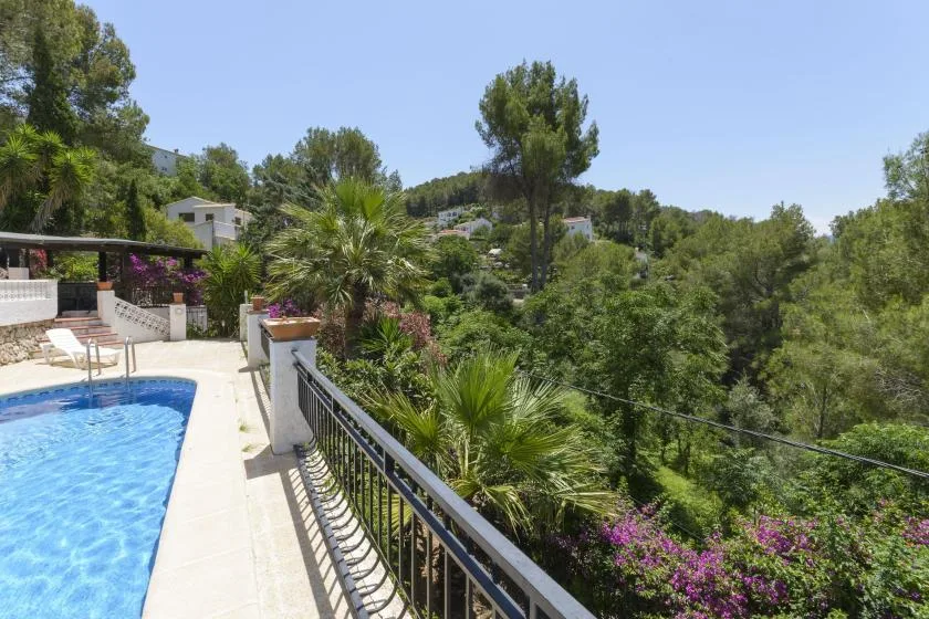 An outdoor pool beside a terrace with railings and a green hillside view.