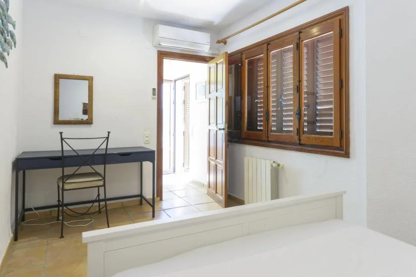 A bright bedroom with a desk, open doorway, and wooden window shutters.