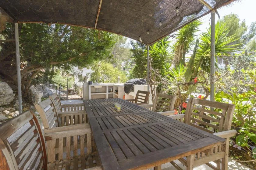 An outdoor dining area has a long wooden table and seating beneath a shade canopy.