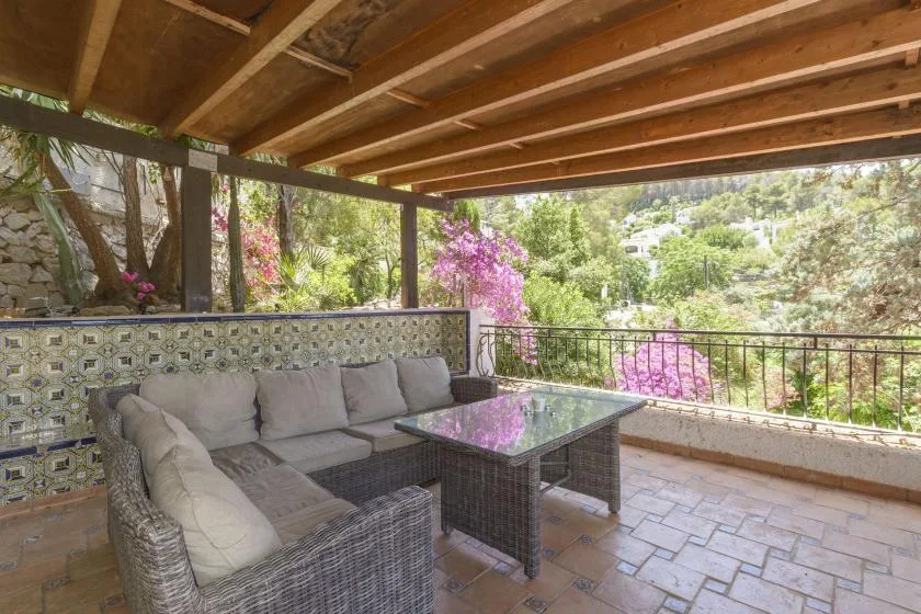 A covered outdoor seating area with a glass-topped table overlooks a garden.