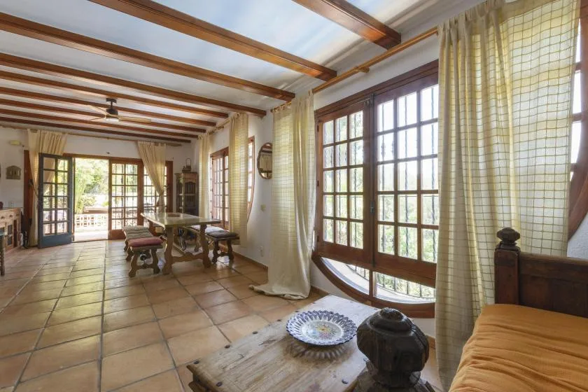 A sunlit living room with large windows, tiled floors, and wooden ceiling beams.