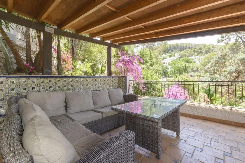 A covered outdoor seating area with a sectional and glass-top table overlooks a leafy garden.