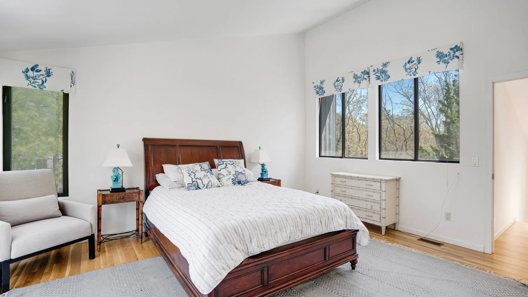Un dormitorio con una cama de madera, ropa de cama blanca y ventanas con vista a los árboles.