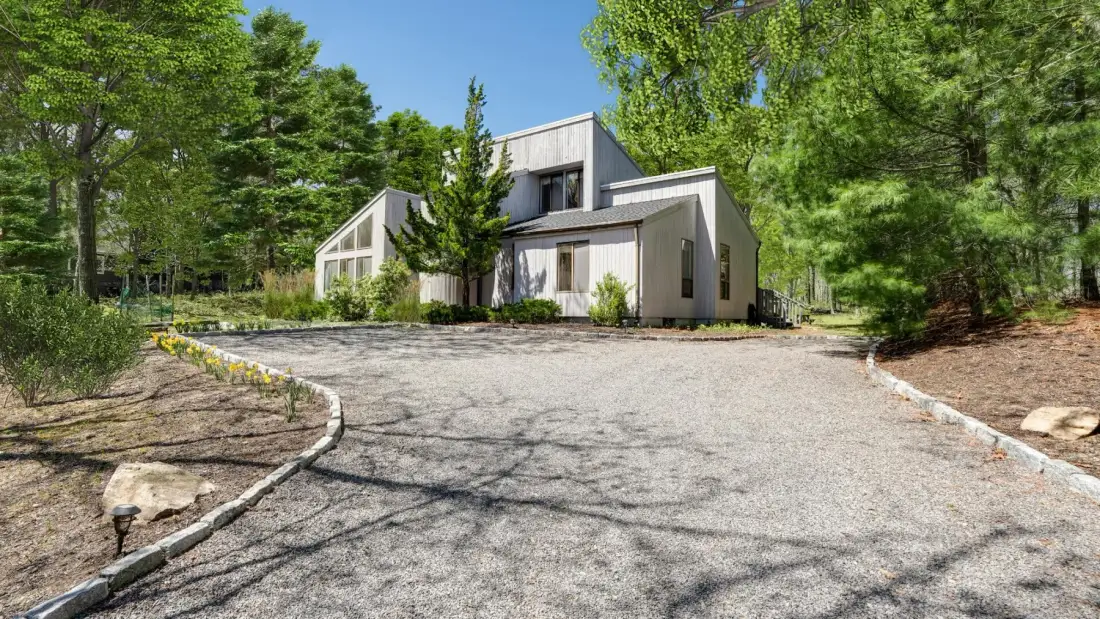 Una casa moderna se encuentra al final de una entrada pavimentada, rodeada de árboles.