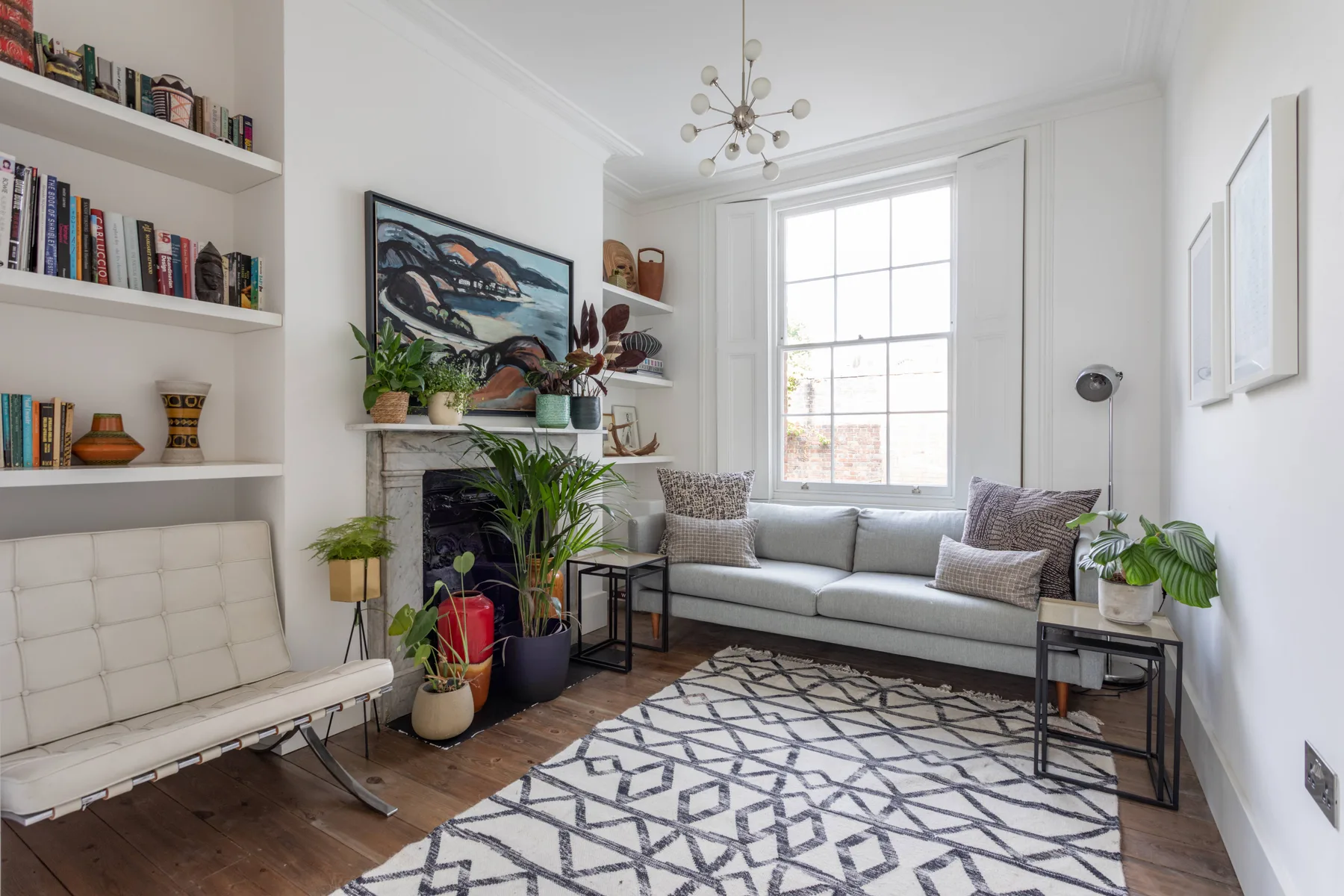 Una sala luminosa con un sofá gris, una ventana grande y plantas frondosas.