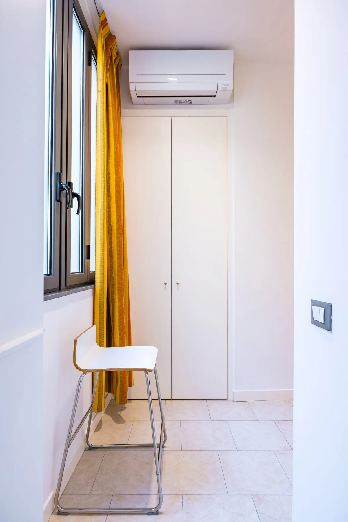 Una esquina luminosa con una ventana, cortinas amarillas, una silla blanca y un aire acondicionado de pared.