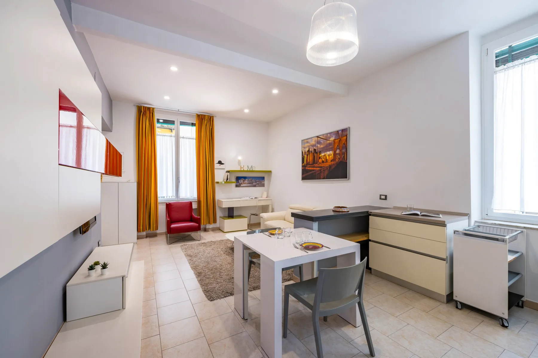 Un estudio luminoso con cocina pequeña, mesa de comedor y ventana con cortinas naranjas.
