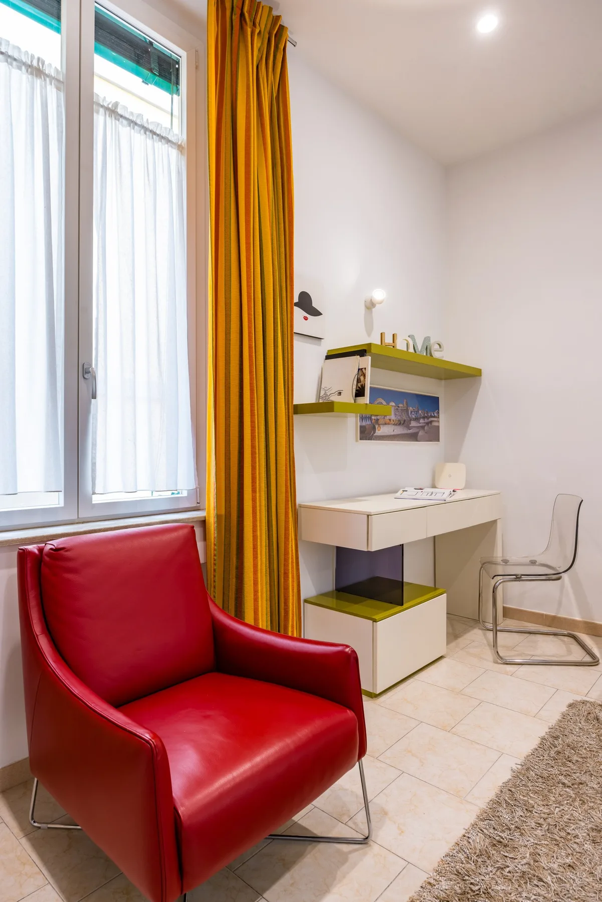 Un cuarto luminoso con un sillón rojo junto a una ventana con cortinas y un escritorio pequeño.