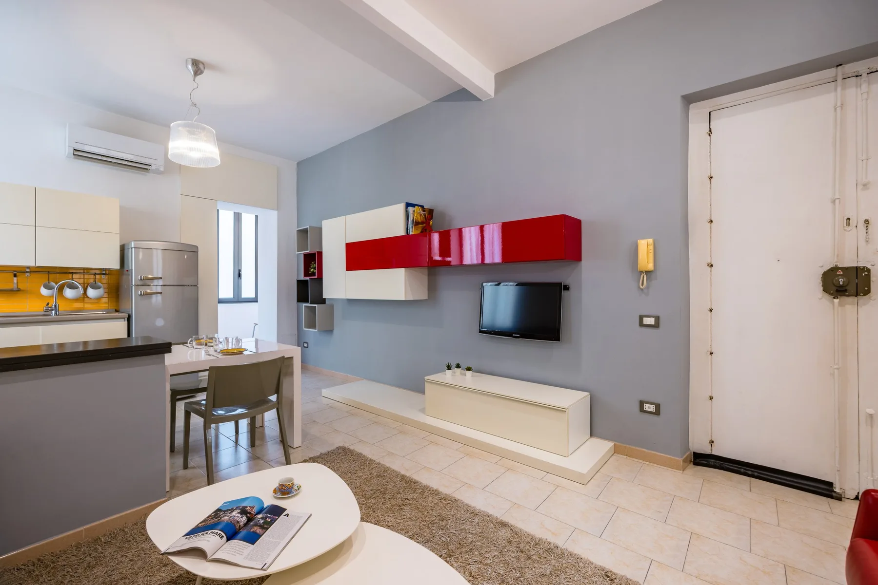 Una sala de estar con televisor en la pared y estantes rojos junto a un comedor.