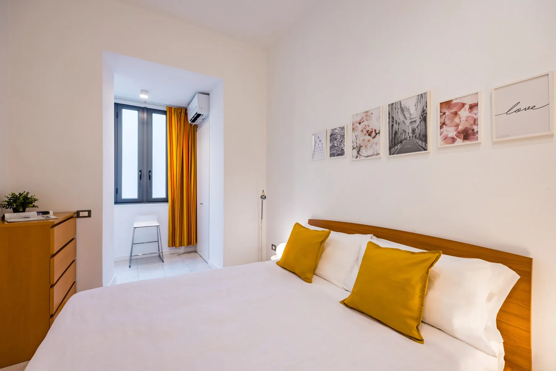 Un dormitorio con una cama, cojines color mostaza y cuadros enmarcados sobre el cabecero.
