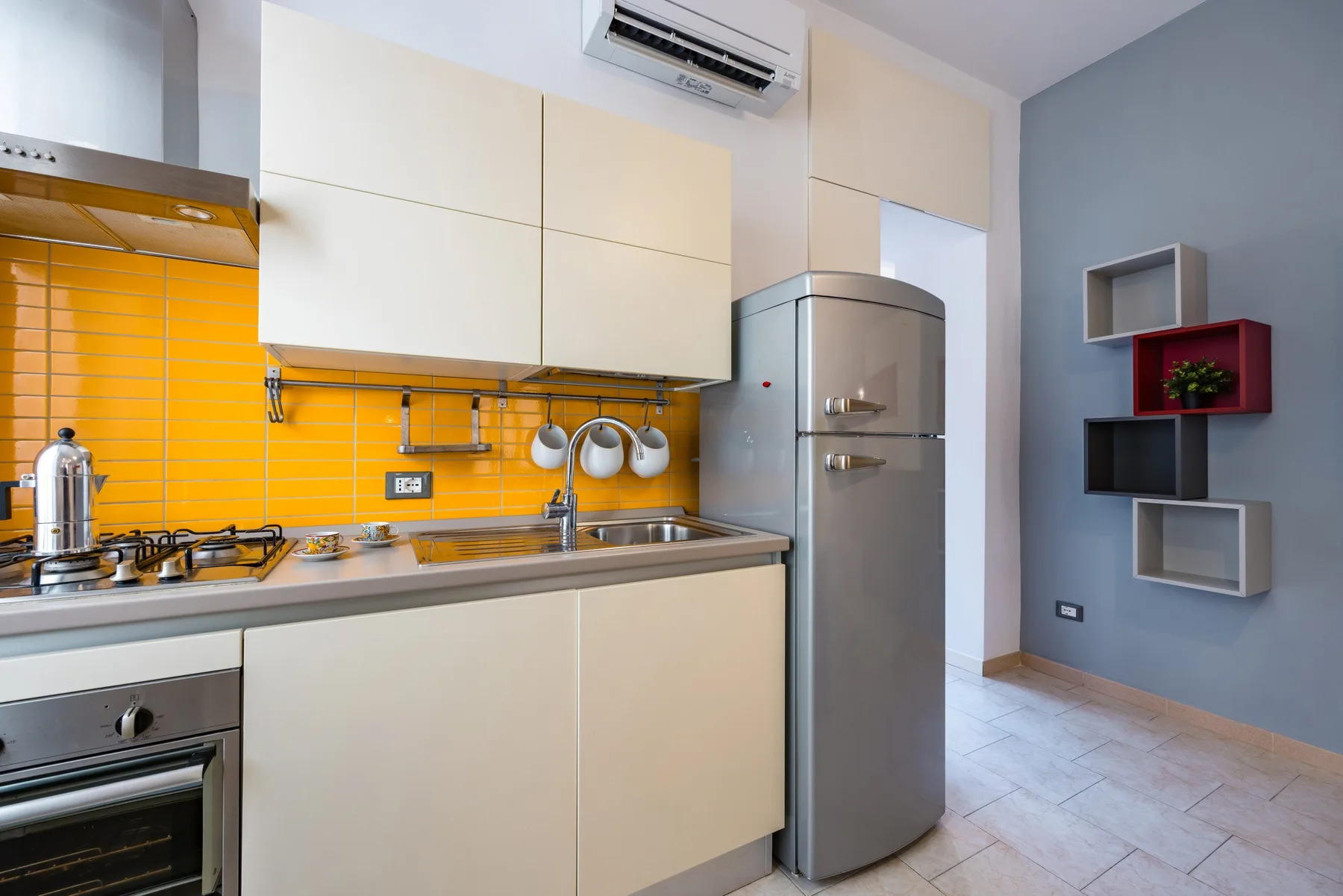Una cocina compacta con armarios color crema, un refrigerador de acero inoxidable y un salpicadero naranja.