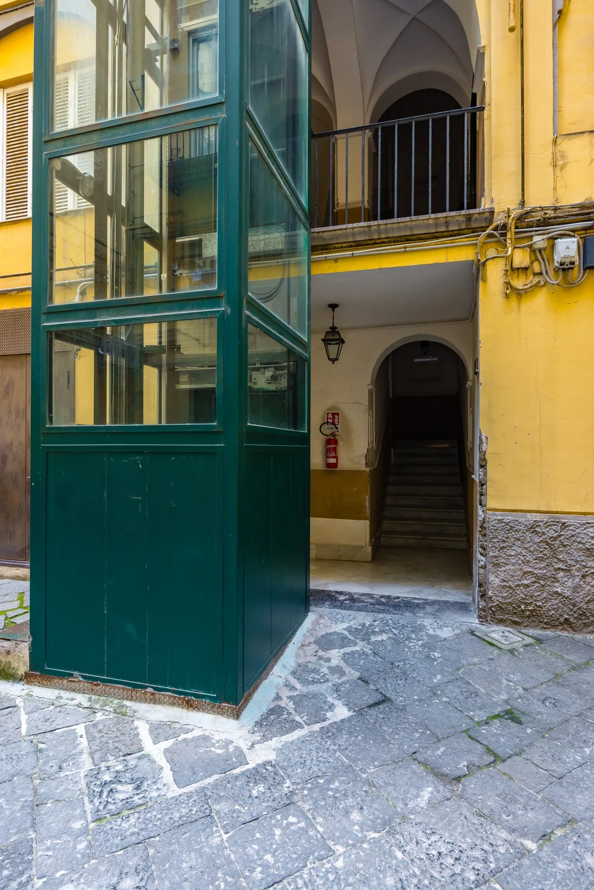 Un ascensor verde de vidrio está junto a una entrada arqueada en un edificio amarillo.