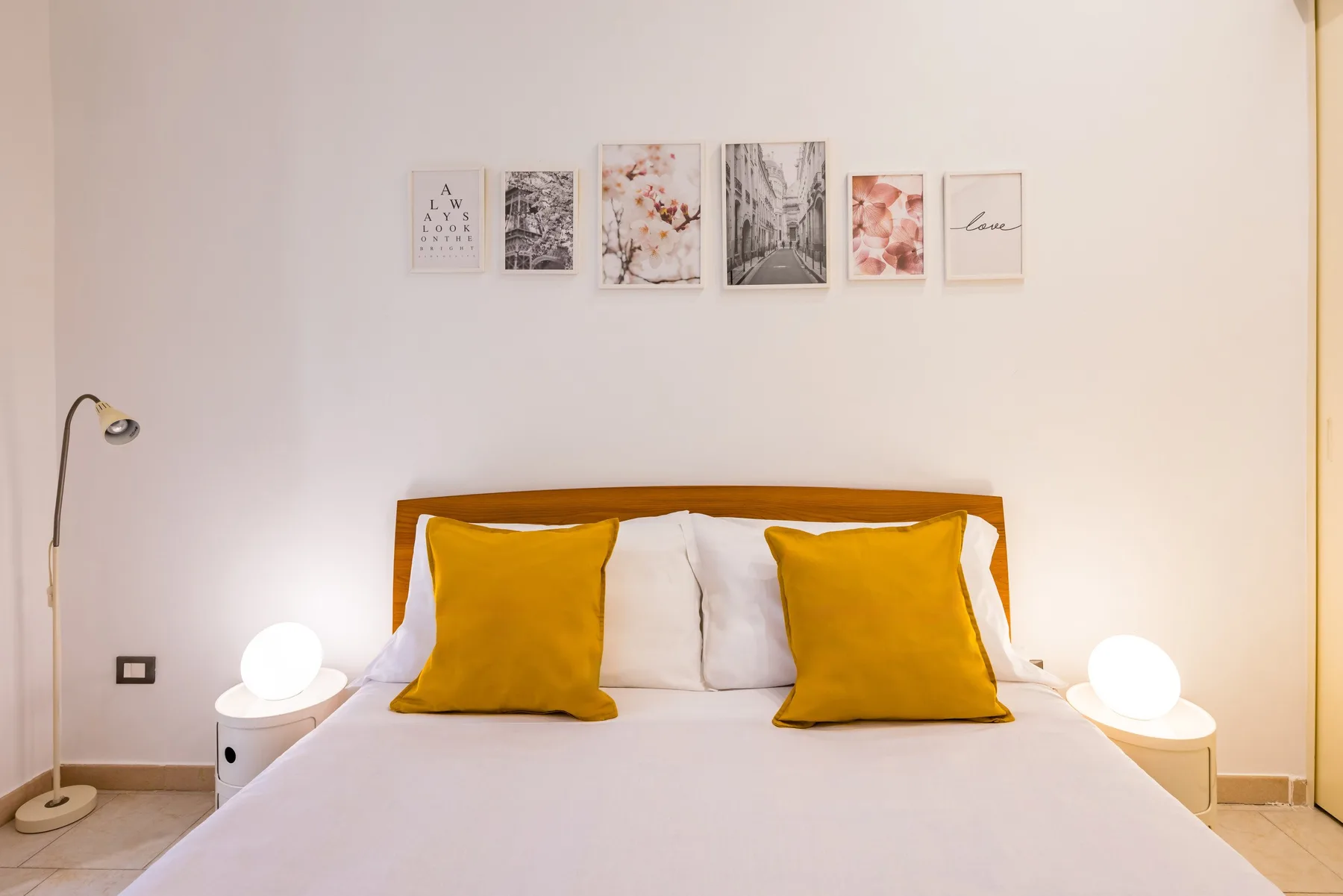 Un dormitorio con una cama, almohadas color mostaza y láminas enmarcadas sobre el cabecero.