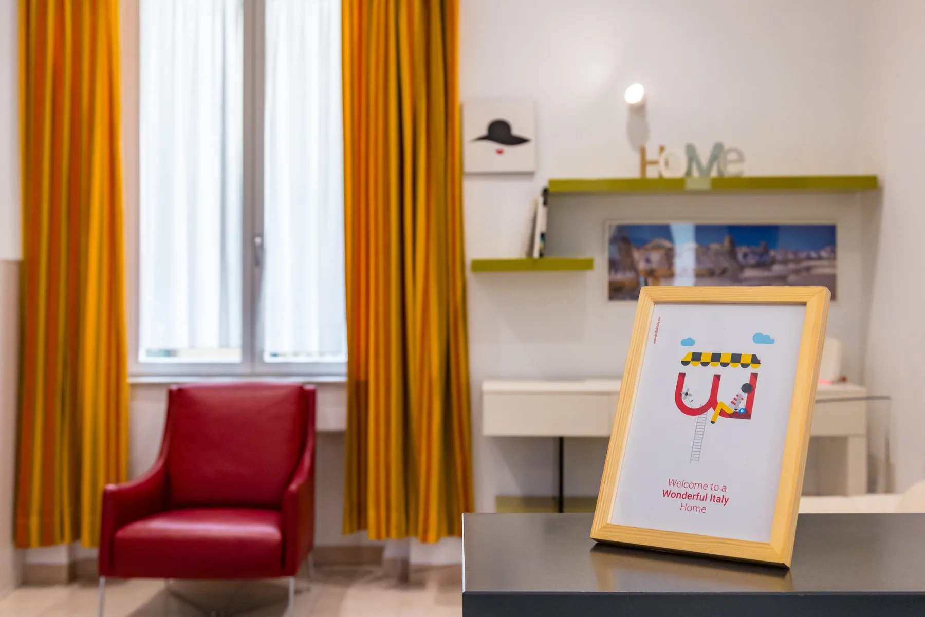 Una habitación con un sillón rojo, cortinas de colores vivos y un cartel de bienvenida enmarcado.