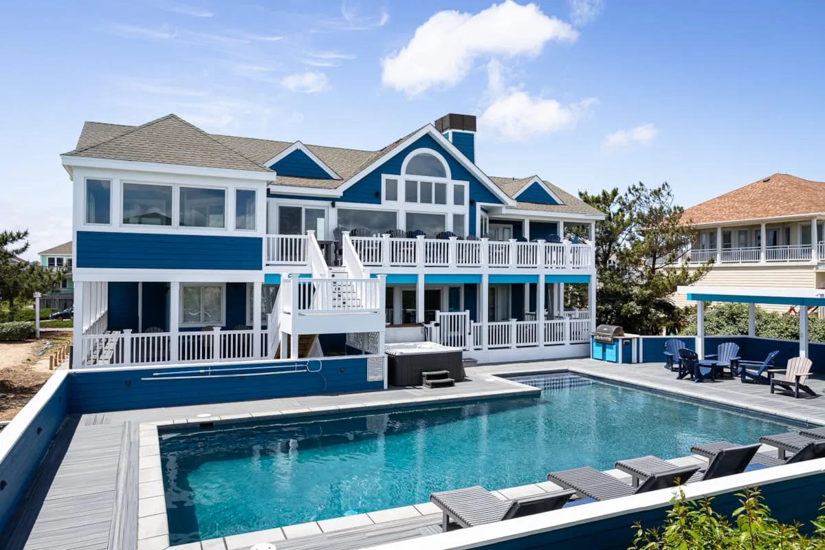 A large blue-and-white house stands beside a swimming pool and outdoor seating.