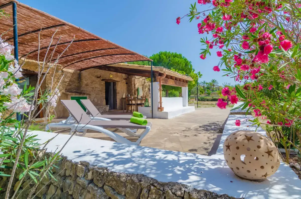 A stone patio with lounge chairs sits beneath a shaded terrace beside flowering shrubs.