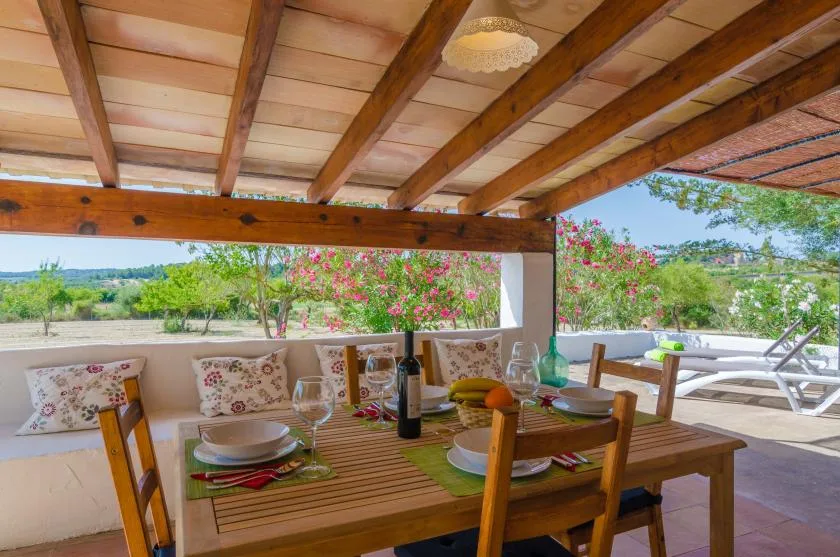 A covered outdoor dining area with a wooden table and chairs overlooks a garden.