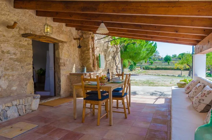 A covered stone patio with a wooden dining table overlooking a green landscape.