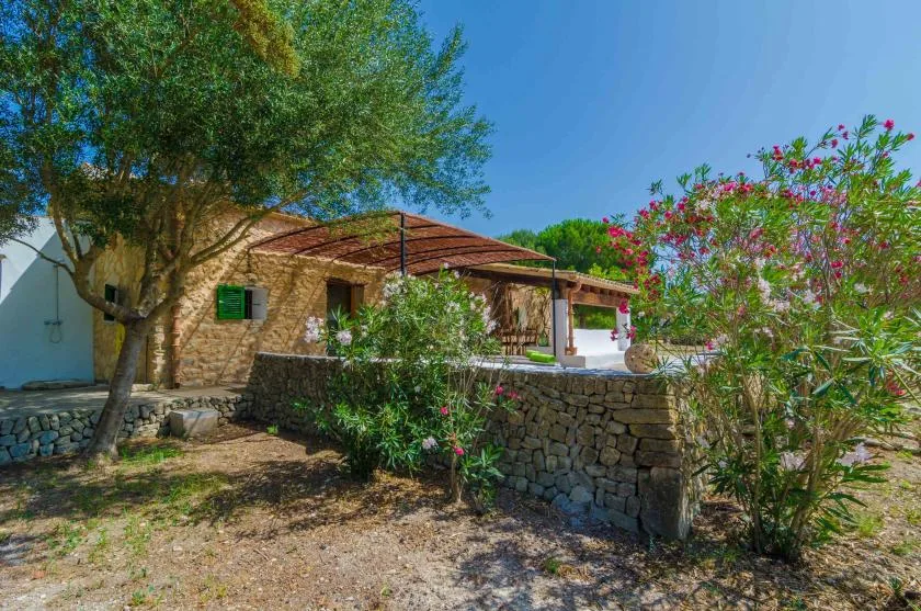 A stone house sits behind a low stone wall, with trees and flowering shrubs around it.