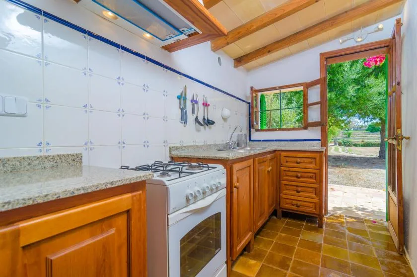 A tiled kitchen with wooden cabinets, a stove, and an open door to greenery.