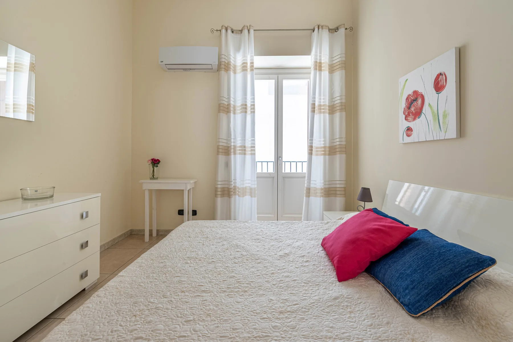 Un dormitorio luminoso con una cama, cojines de colores y ventanas altas con cortinas.