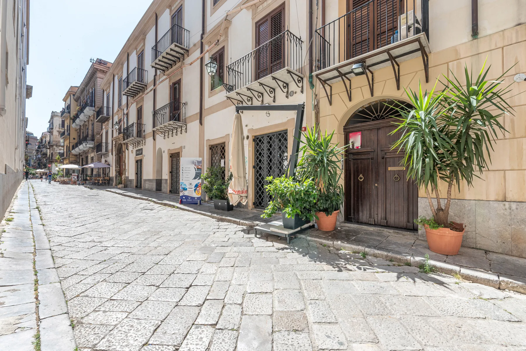 A sunlit cobblestone street lined with buildings and potted plants.