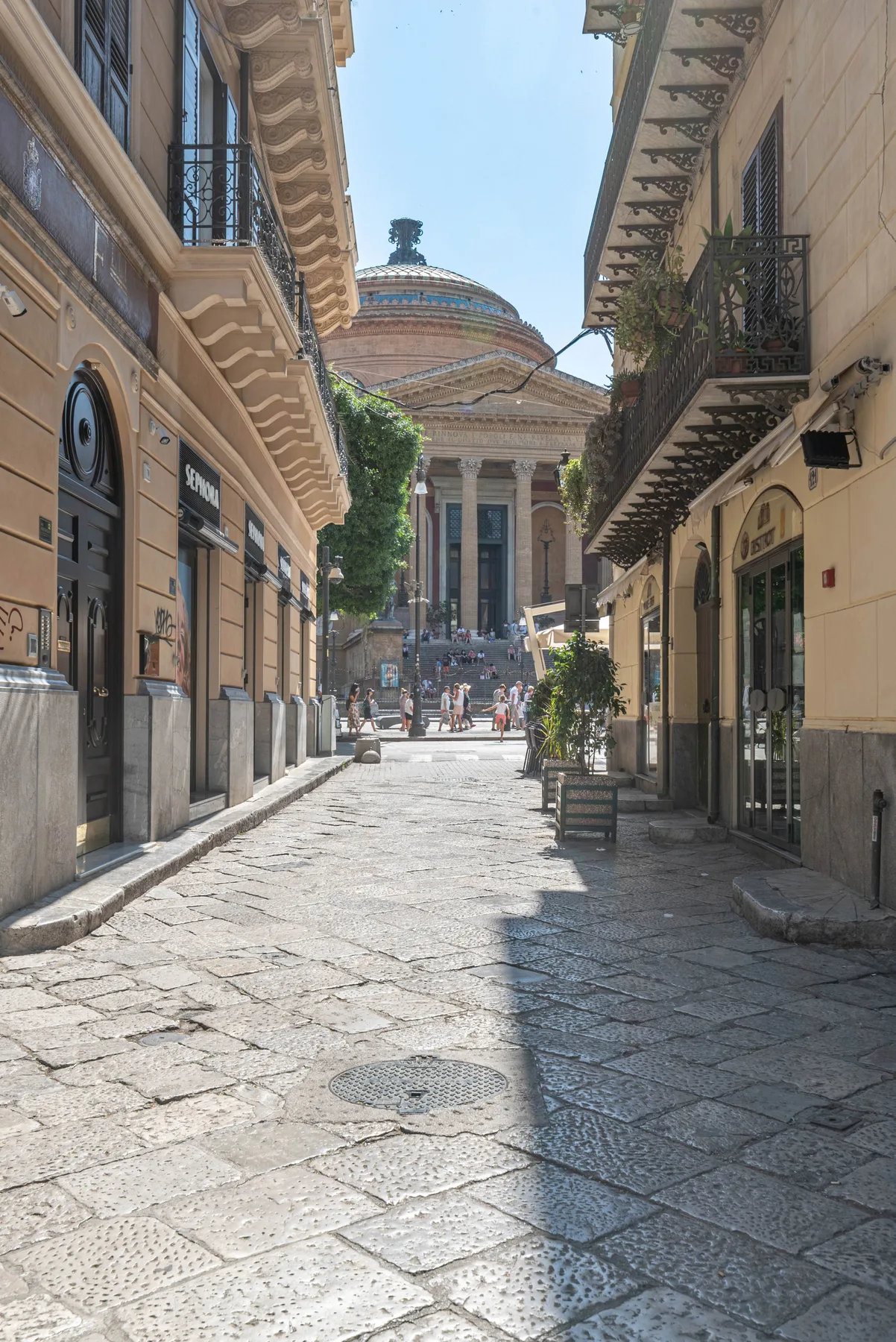 A narrow stone-paved street leads toward a domed building between tall façades.