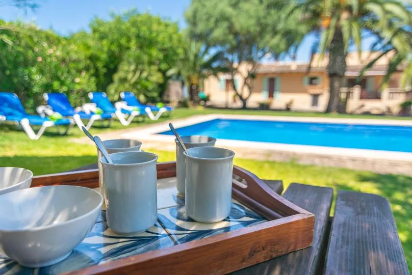 A poolside table holds cups and bowls, with lounge seating and a swimming pool beyond.
