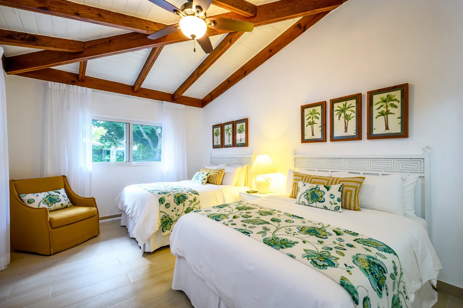 Un dormitorio luminoso tiene dos camas con ropa de cama blanca y estampada en verde bajo vigas de madera.