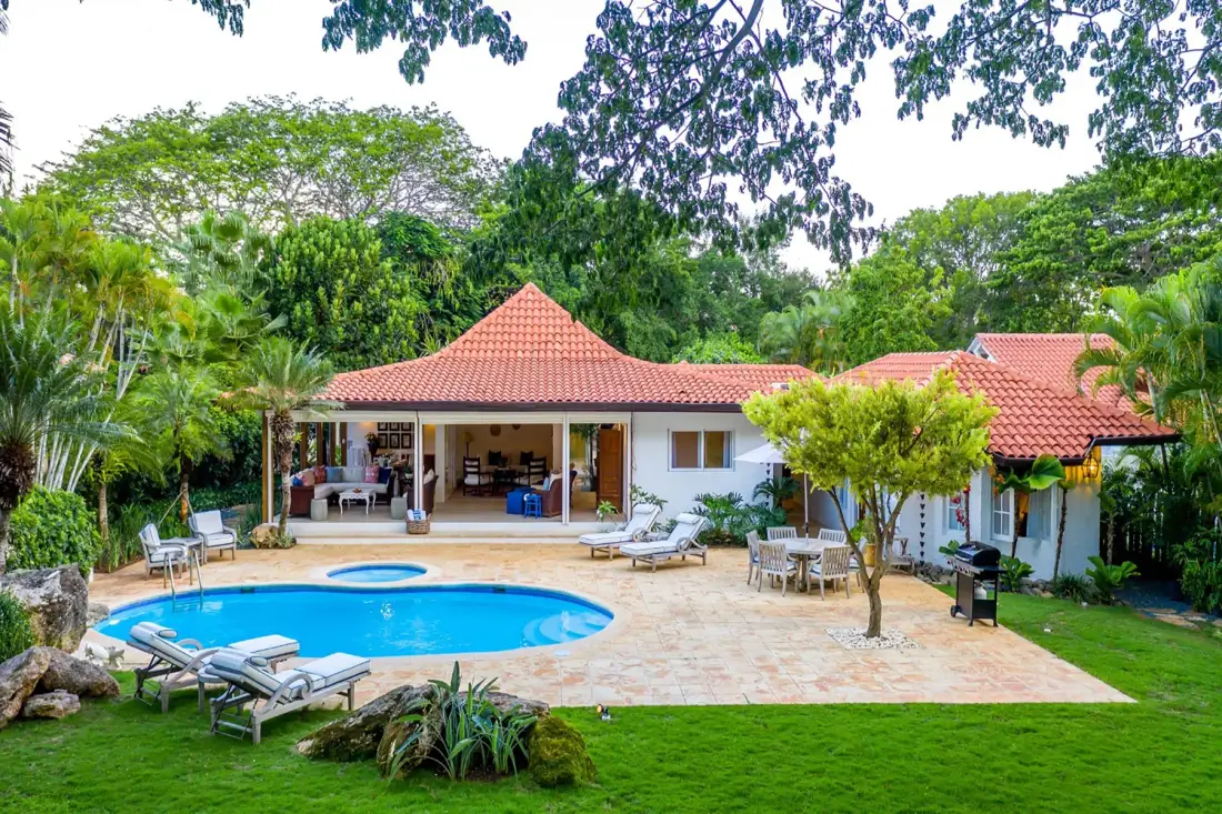 Una casa con techo de tejas rojas da a una piscina y un patio pavimentado rodeados de vegetación.
