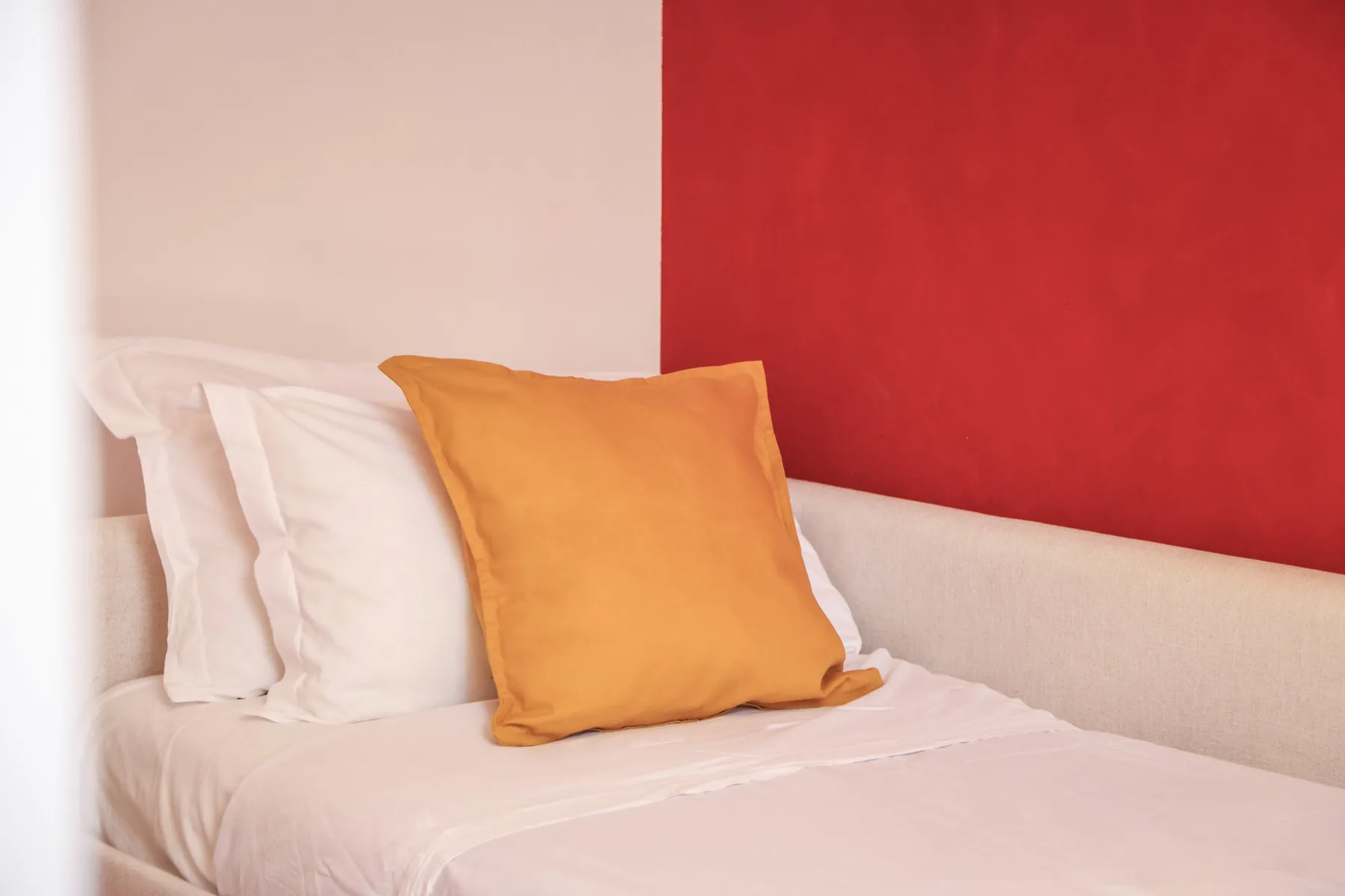 A bed with white bedding and an orange accent pillow against a red wall.