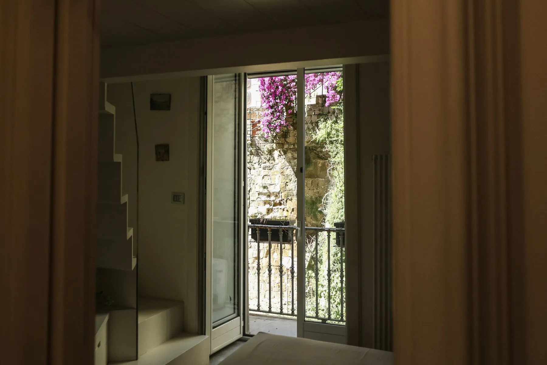 A dim interior opens onto a glass balcony door with greenery outside.