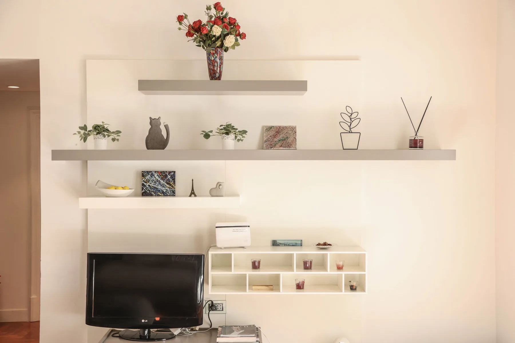A bright living room features a television beneath white wall shelves with decorative objects.