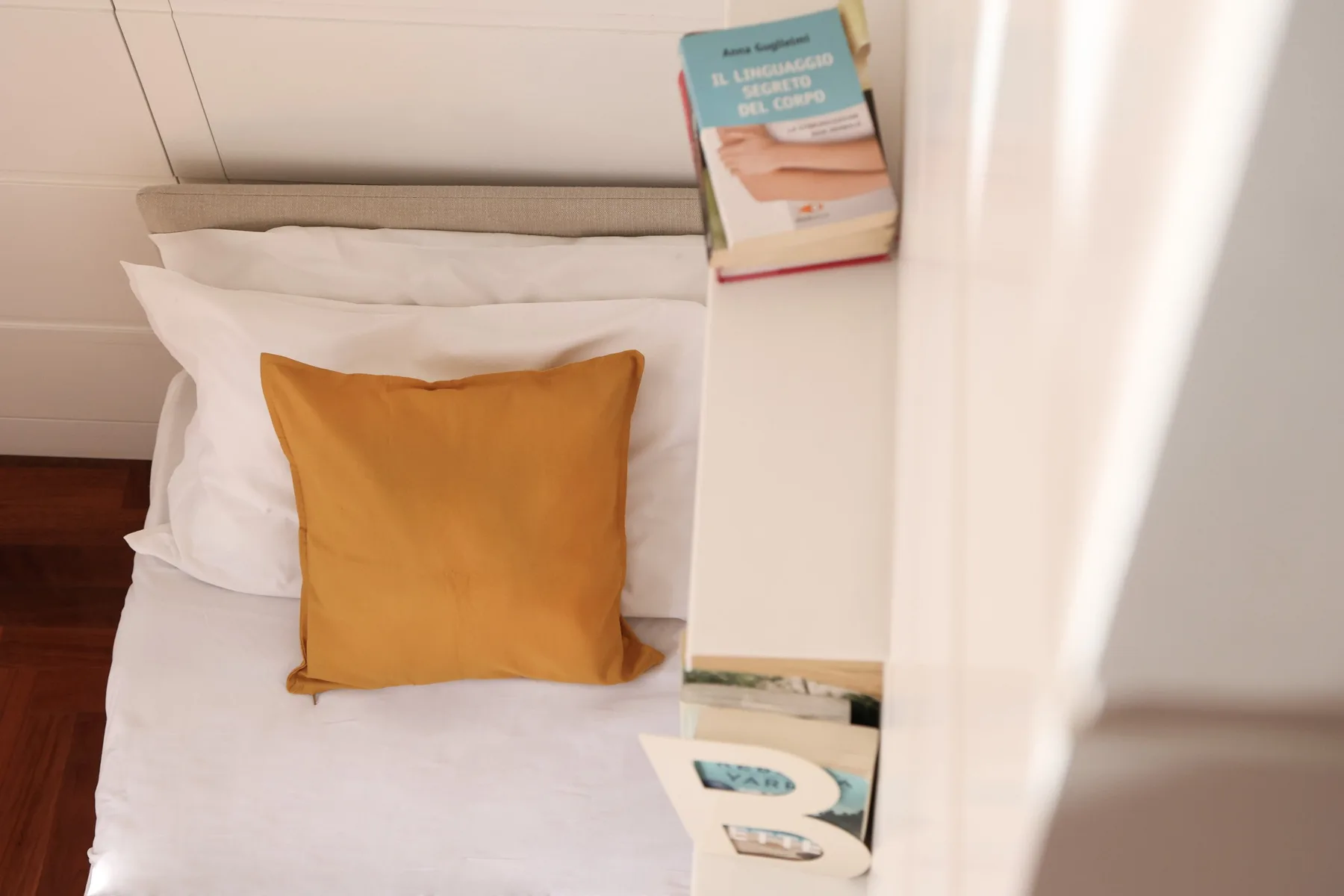 A bed with white bedding and an orange accent pillow beside a narrow shelf.