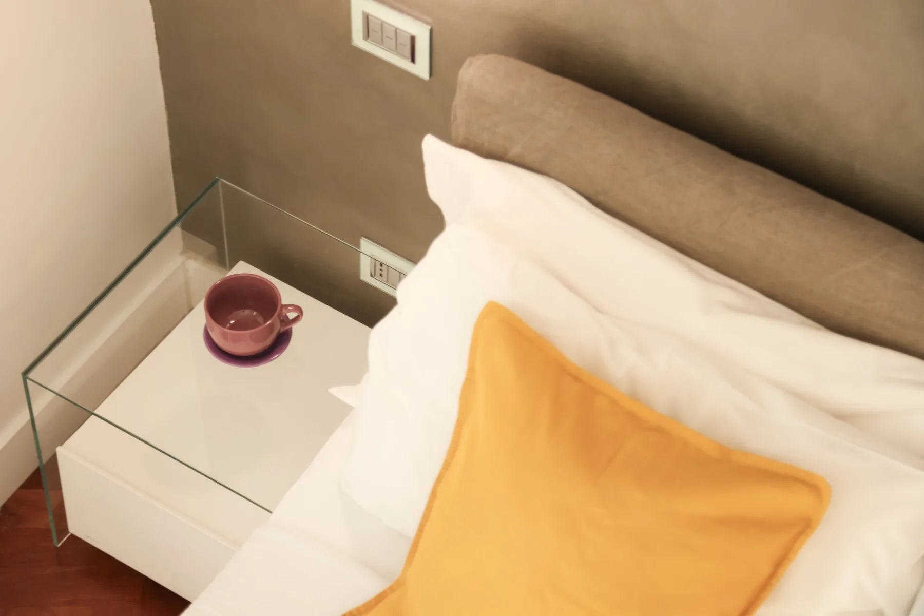 A bedroom bed with white bedding, a yellow cushion, and a glass bedside table.