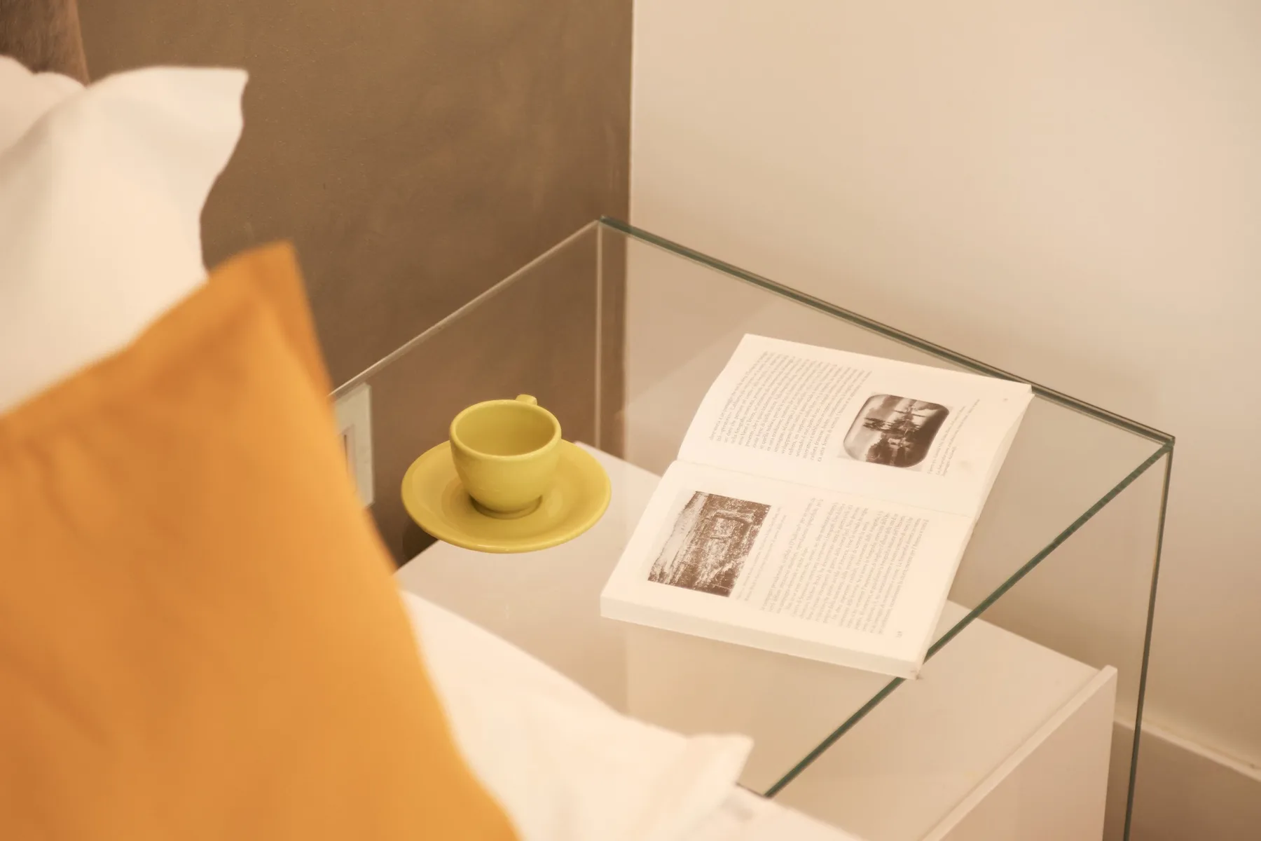 A glass bedside table holds an open book and a yellow cup and saucer.