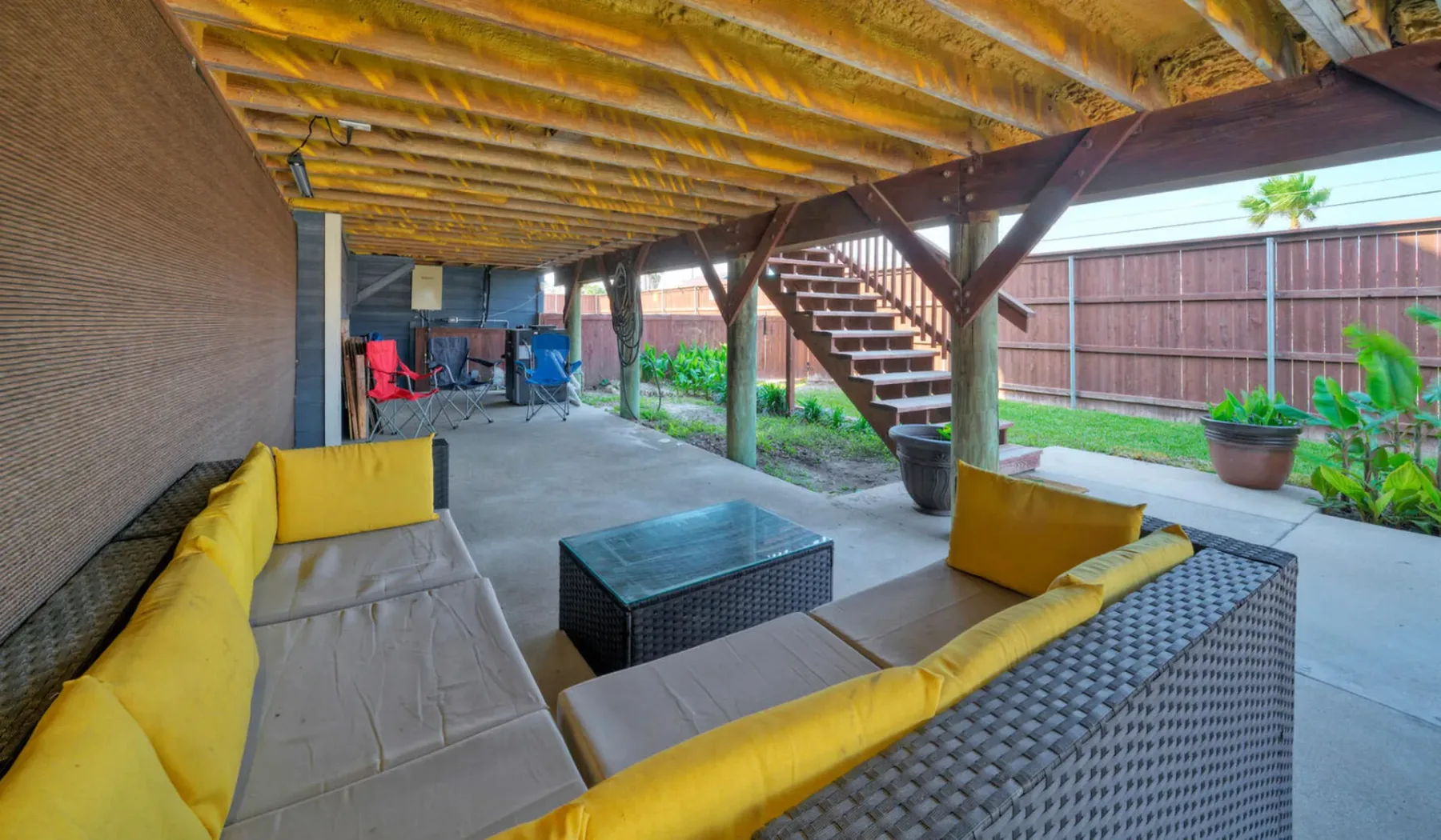 Un patio cubierto con asientos al aire libre y una escalera junto a un jardín cercado.