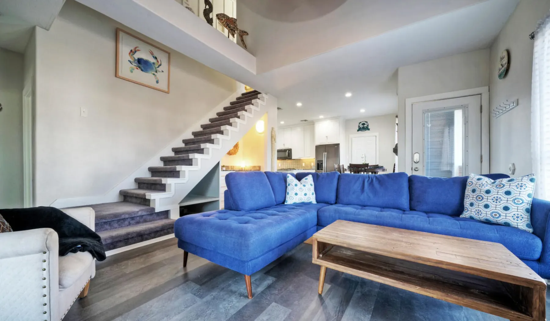 Una sala luminosa con asientos azules y una mesa de centro de madera bajo una escalera.