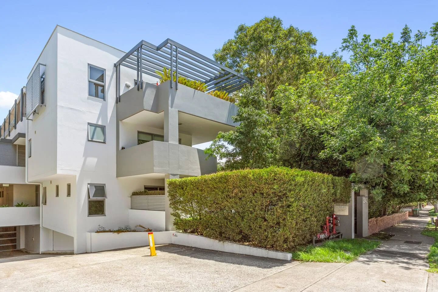 A modern white building stands beside a leafy sidewalk and trimmed hedge.