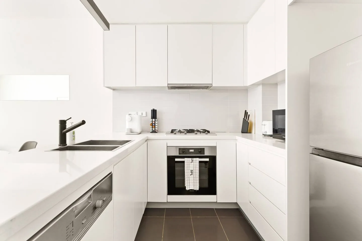 Una cocina blanca y luminosa tiene un horno debajo de la placa y un fregadero junto a la encimera.