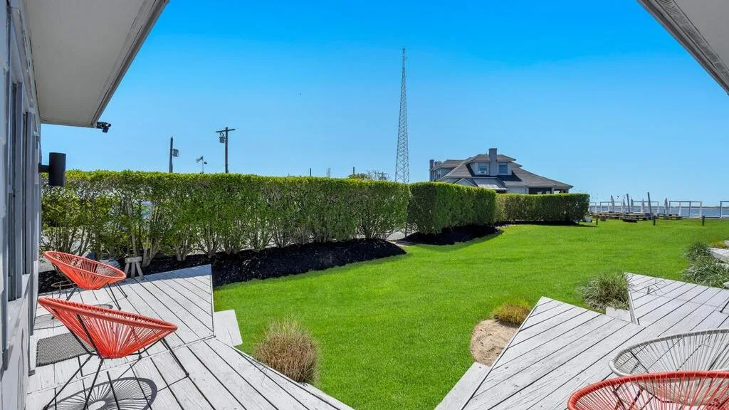 Un patio exterior con césped y setos, junto a una terraza con asientos rojos.