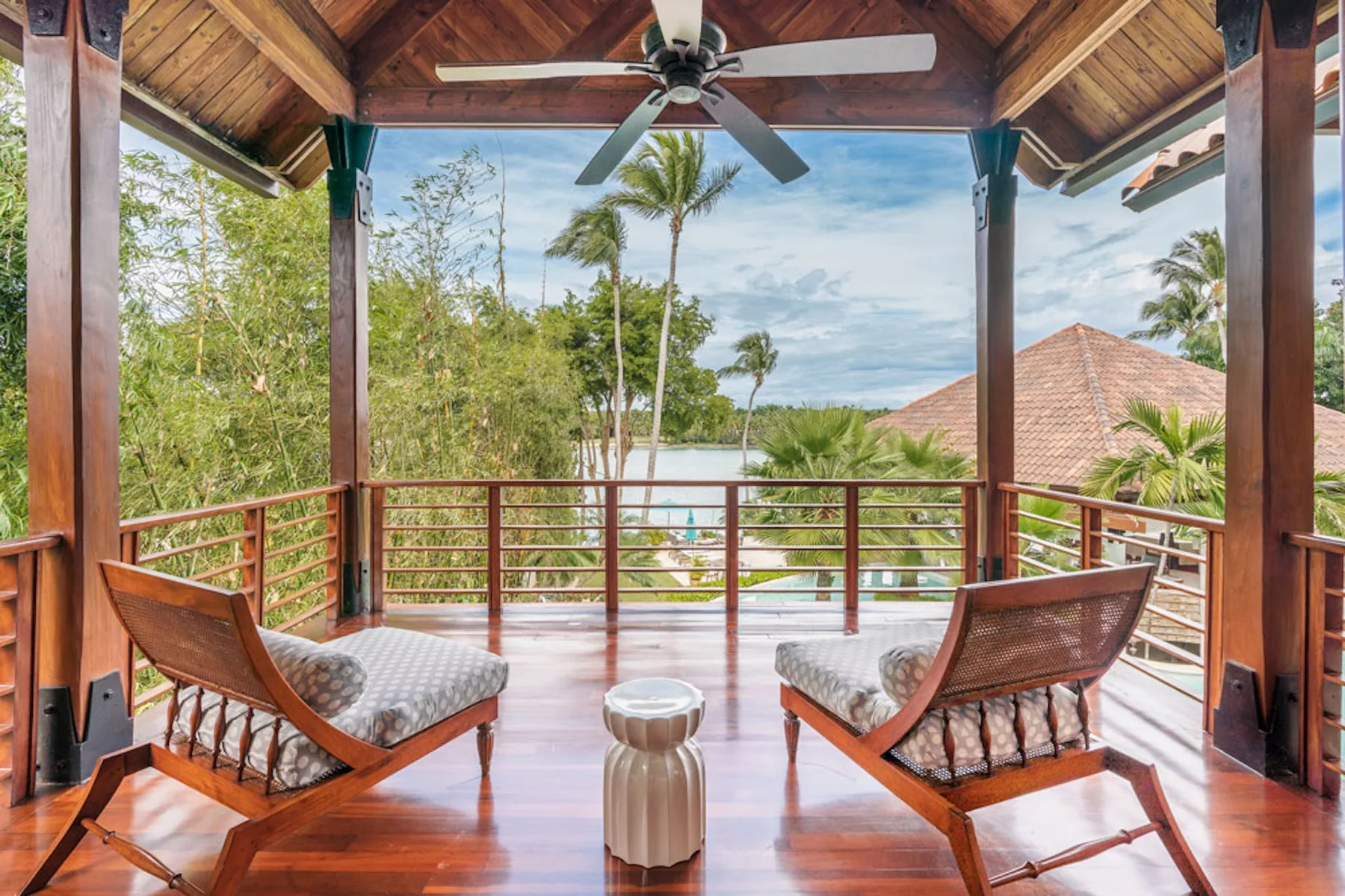 A covered wooden balcony has two cushioned lounge chairs facing a leafy view.