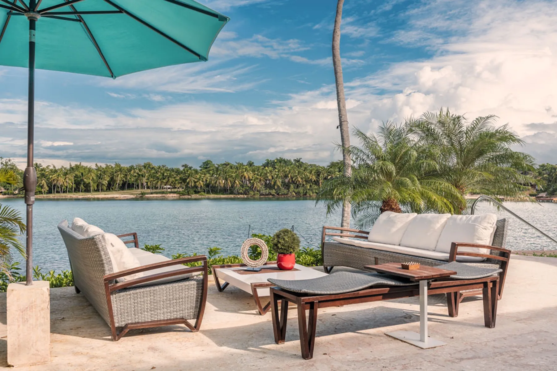 Outdoor seating beside a calm waterfront, with a teal umbrella overhead.