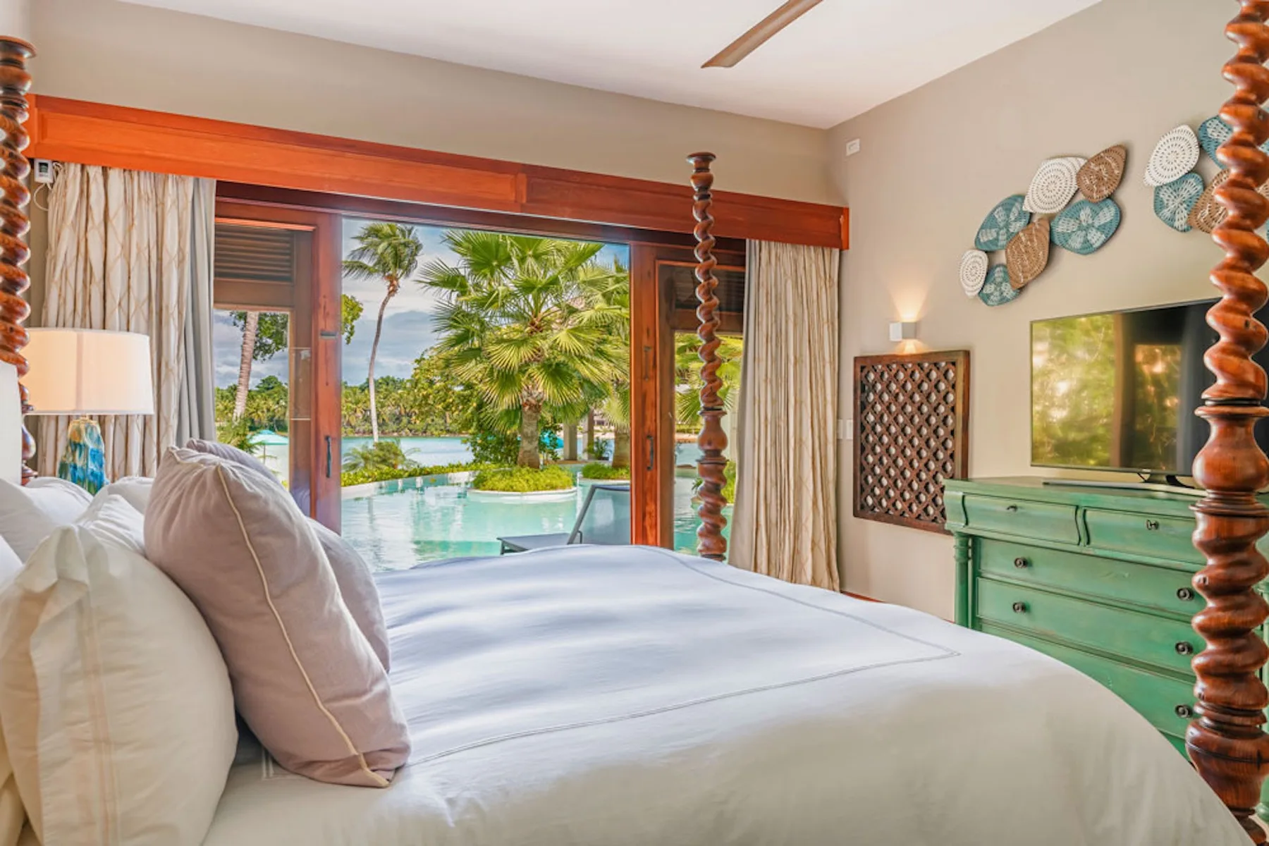 A bedroom with a bed facing open doors and a view of a pool and palm trees.