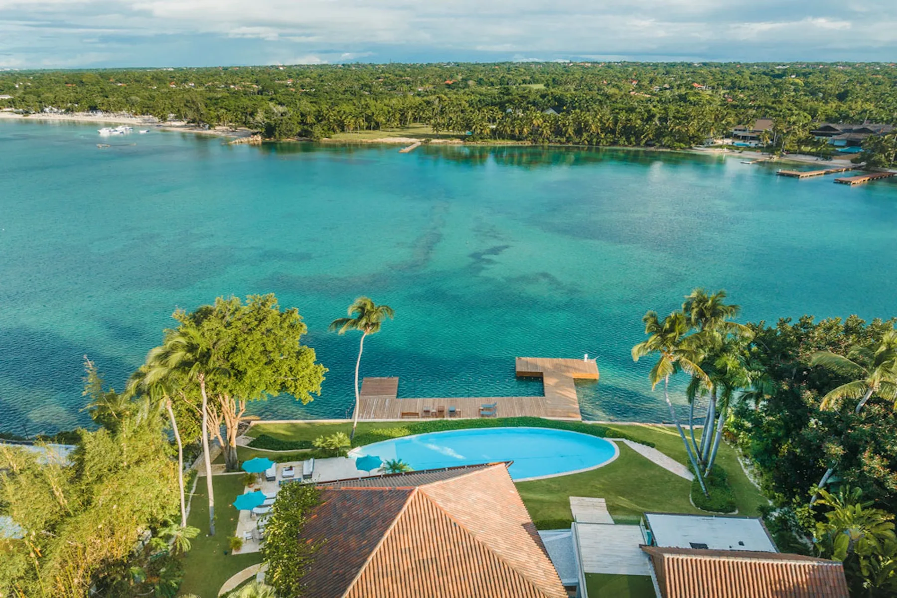 An aerial view shows a coastal property beside turquoise water, with a pool and palm trees.