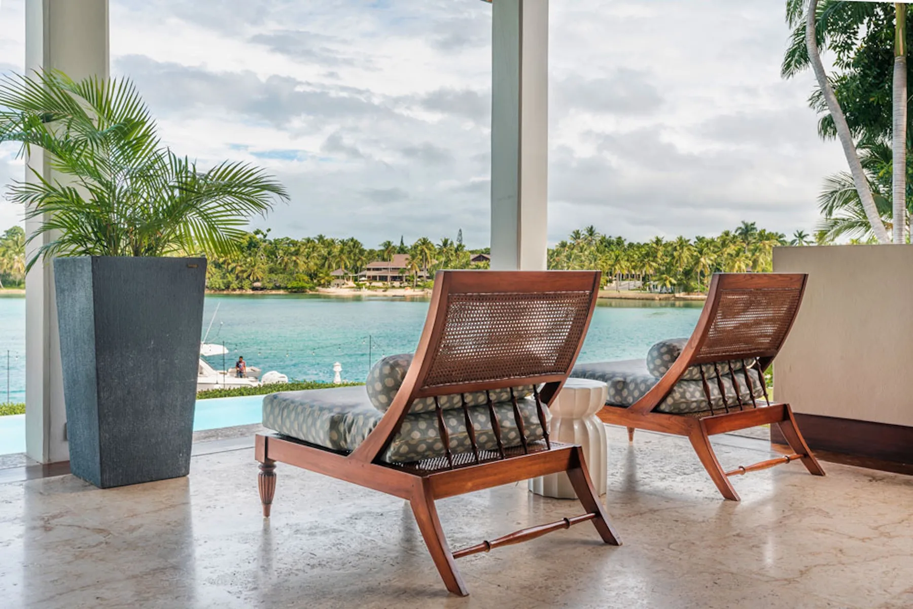 A covered patio overlooks a pool and tropical water, with two lounge chairs.